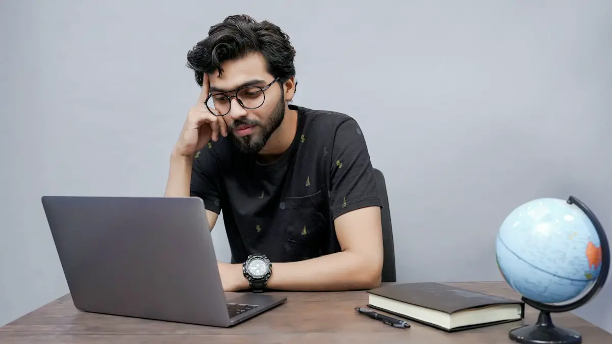 A student at a desk thinking over a question on his laptop, with a notebook beside him.
