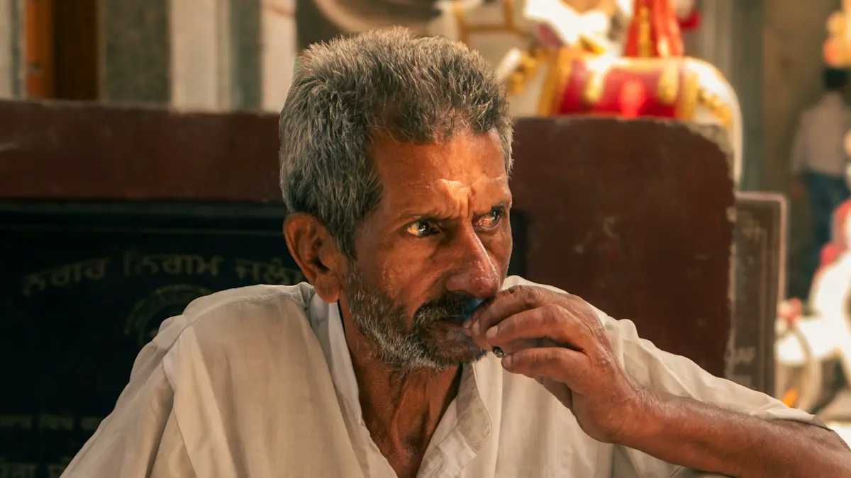 Older man with beard and tilak sits, hand to mouth.